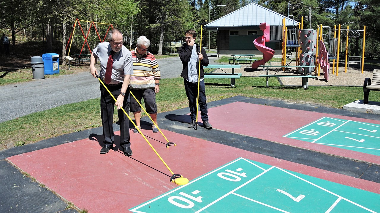 De gauche à droite : S'amusant sur les terrains de shuffleboard, Claude Morin, maire de la Ville de Saint-Georges, Carole Paquet, directrice du Service des loisirs et de la culture de la Ville de Saint-Georges, et Samuel Duval, stagiaire au Service des loisirs et de la culture de la Ville de Saint-Georges, De gauche à droite : Carole Paquet, directrice du Service des loisirs et de la culture de la Ville de Saint-Georges, Claude Morin, maire de la Ville de Saint-Georges, et Samuel Duval, stagiaire au Service des loisirs et de la culture de la Ville de Saint-Georges, lors de la conférence de presse annonçant la programmation estivale 2018 au parc des Sept-Chutes de Saint-Georges, jeudi le 24 mai 2018.
