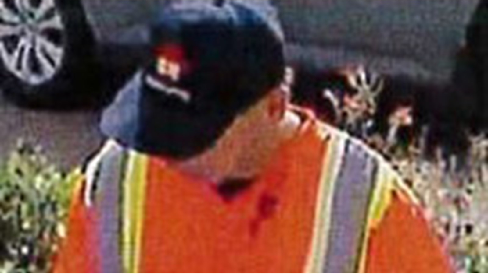 Il porte une casquette marine avec un logo rouge et blanc, une veste orange de travailleur, un pantalon beige et des souliers noirs.