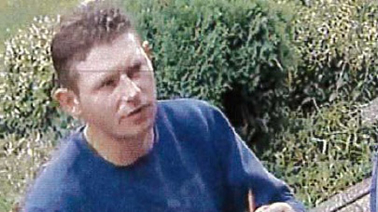 Environ 35 ans, il a les cheveux bruns. Il porte un chandail à manches longues bleu, un pantalon noir et des souliers gris.